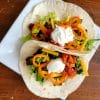 Two finished soft shell tacos with sliced peppers, shredded cheese and a dallop of sour cream on a square white plate.