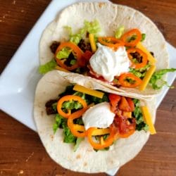 Two finished soft shell tacos with sliced peppers, shredded cheese and a dallop of sour cream on a square white plate.