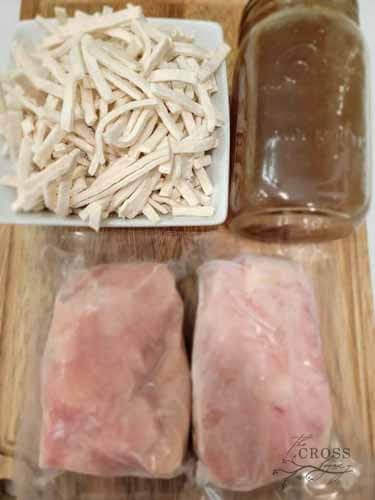 Dish of noodles, jar of chicken stock and package of frozen chicken on a wooden cutting board. Ready to make homemade chicken noodle soup.