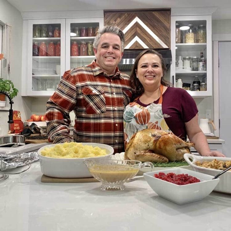 Mike and Amy Cross side-by-side in their kitchen on Thanksgiving with their beautifully roasted turkey, mashed potatoes, gravy, cranberry relish, and stuffing in front of them.