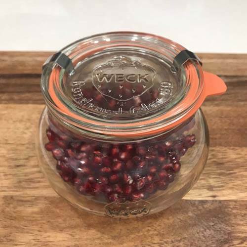 Pomegranate seeds in a glass jar, sitting on a wooden cutting board.