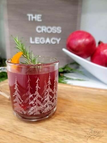 Holiday drink with a sprig of Rosemary and a wedge of orange sitting on a wooden cutting board with a bowl of Pomegranates and a The Cross legacy sign behind it.
