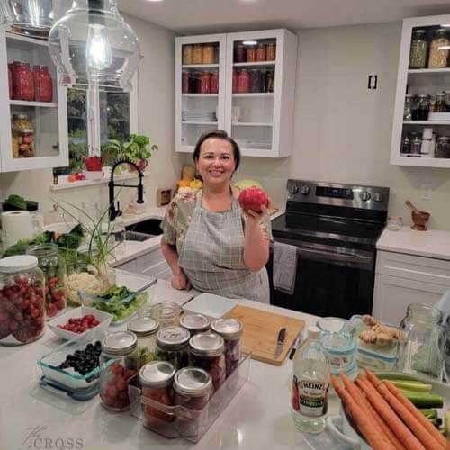 Amy in her kitchen holding a pomegranate with a spread of produce in jars and glass containers on the kitchen counter around her.