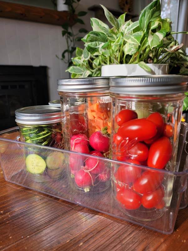 Sliced cucumbers, radishes, and cherry tomatoes in glass mason jars in a salad basket on a wooden table.