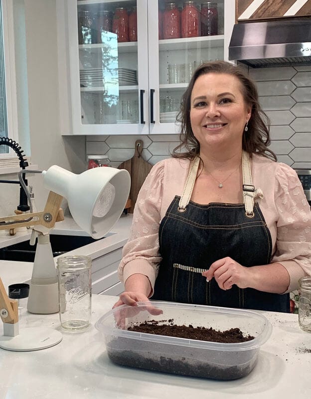 Amy Cross standing at her kitchen counter with all the supplies needed to grow microgreens - a shallow tray, potting soil, seeds, a grow light, and a misting spray bottle.