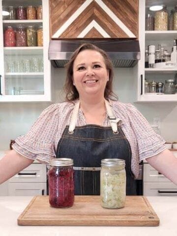 Amy Cross in her kitchen with 2 jars of fermented homemade sauerkraut sitting on a wooden cutting board in front of her.