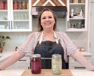 Amy Cross in her kitchen with 2 jars of fermented homemade sauerkraut sitting on a wooden cutting board in front of her.