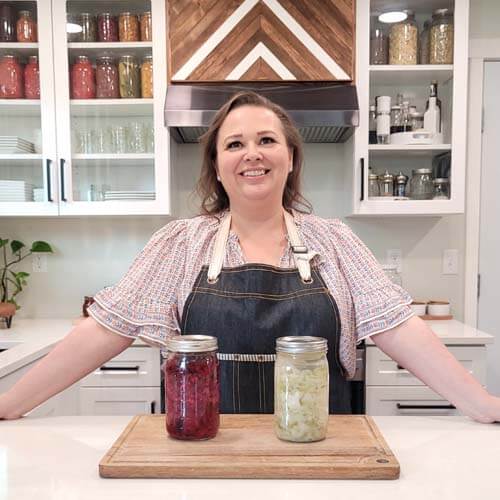 Amy Cross in her kitchen with 2 jars of fermented homemade sauerkraut sitting on a wooden cutting board in front of her.