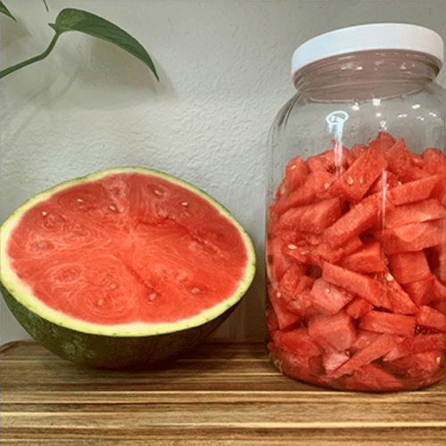 Watermelon-cut-in-half-and-watermelon-cut-into-french-fry-sticks-in-a-gallon-size-jar-sitting-on-a-wood-cutting-board