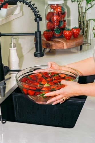 Close up of fresh strawberries soaking in a vinegar bath in a glass bowl. Amy is holding the bowl under the faucet of her kitchen sink.