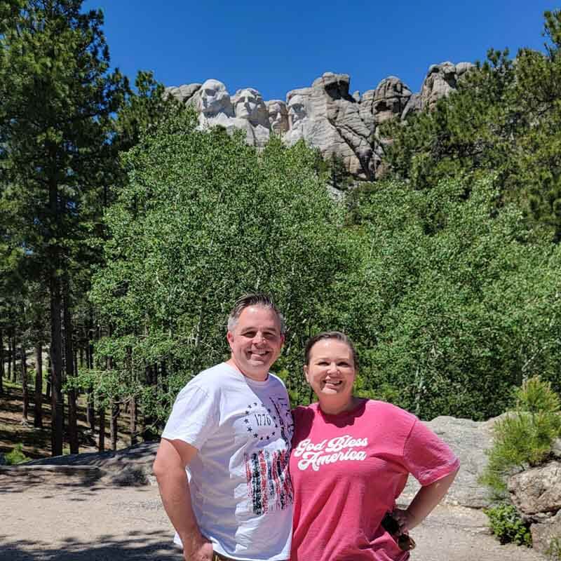mike-and-amy-cross-posing-with-trees-and-Mount-Rushmore-behind-them