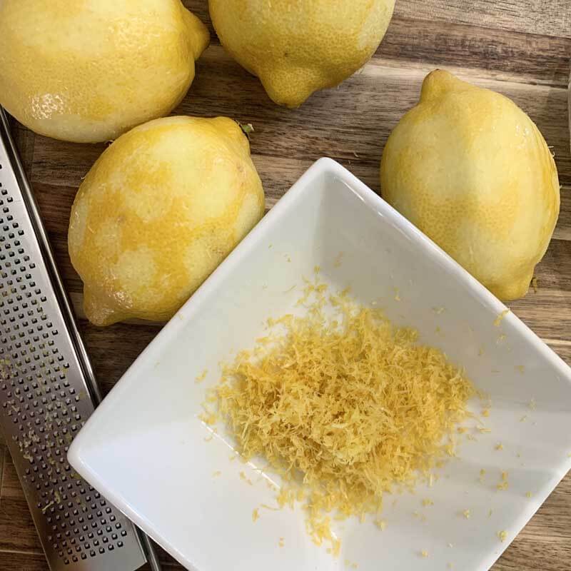 Zested lemons, zester, and small white square dish with lemon zest all on a wooden cutting board