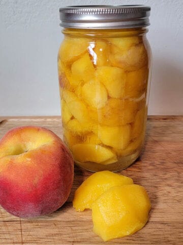 fresh peach and peeled peach used for canning peaches with a jar of canned peaches sitting on a wooden cutting board