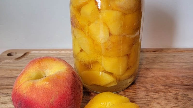 fresh peach and peeled peach used for canning peaches with a jar of canned peaches sitting on a wooden cutting board