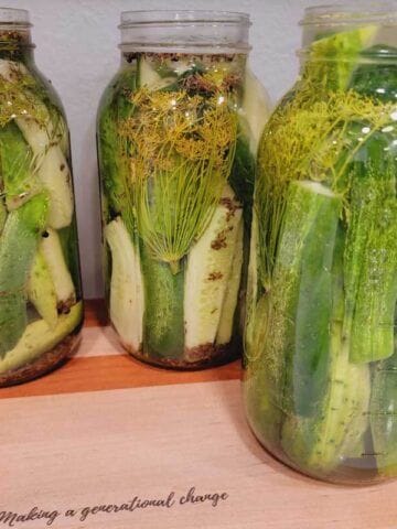 3 jars of finished crunchy dill pickles in jars sitting on a wooden cutting board