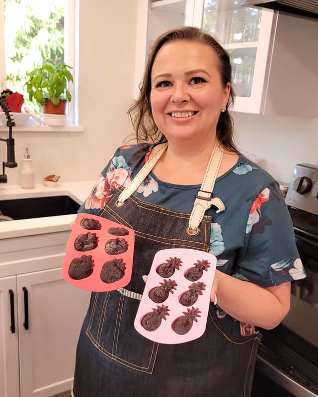 Amy Cross standing in her kitchen holding homemade elderberry gummies in pineapple and strawberry silicone molds