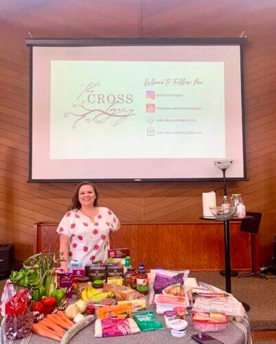 Amy after Step-by-Step event standing at a table with $135 of produce and slideshow behind her
