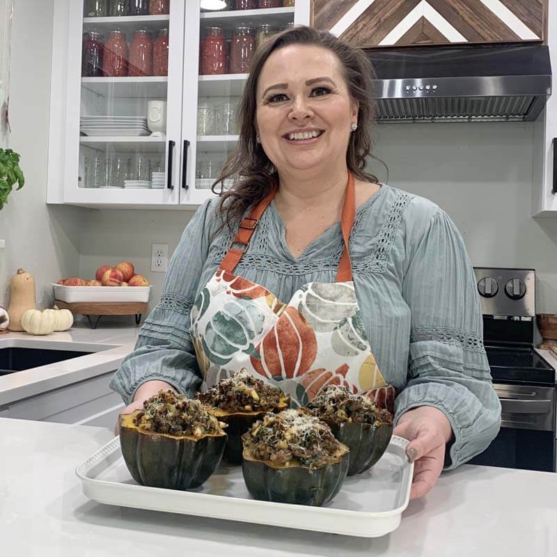 Amy Cross standing at her kitchen counter holding a white tray with four finished stuffed acorn squash.