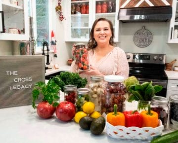 Amy Cross in her kitchen holding a jar of strawberries with a spread of fresh produce on the counter and The Cross Legacy sign. Teaching you how to eat the rainbow on a grocery budget.