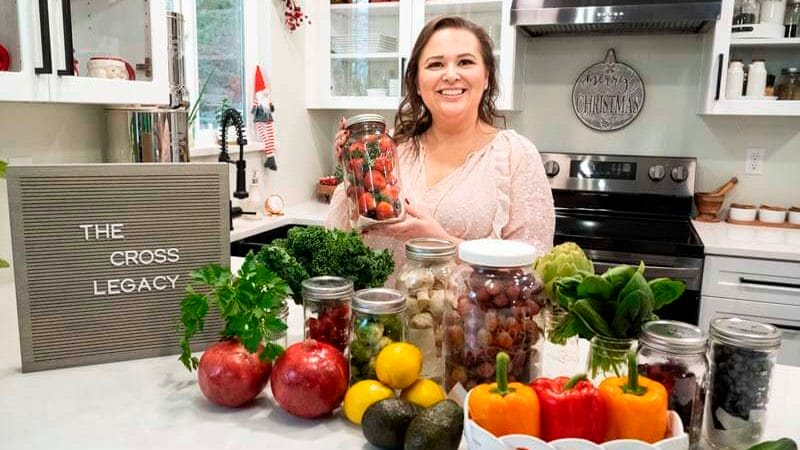 Amy Cross in her kitchen holding a jar of strawberries with a spread of fresh produce on the counter and The Cross Legacy sign. Teaching you how to eat the rainbow on a grocery budget.