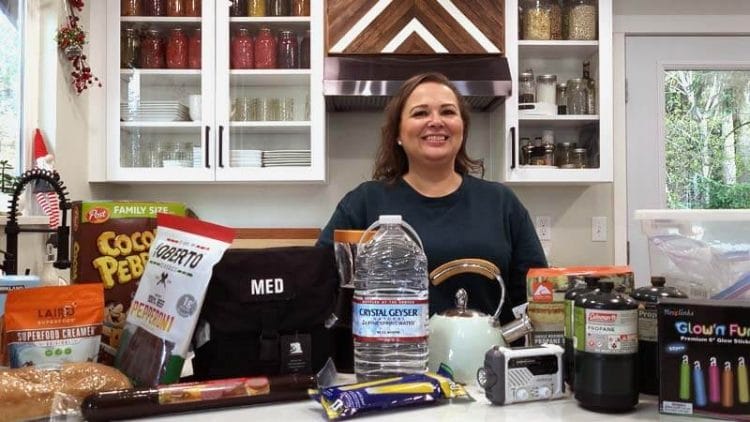 Photo of Amy Cross at her kitchen counter with an array of winter weather emergency planning supplies - glow sticks, bottled water, emergency radio, emergency kit, lantern, shelf-stable foods, etc.