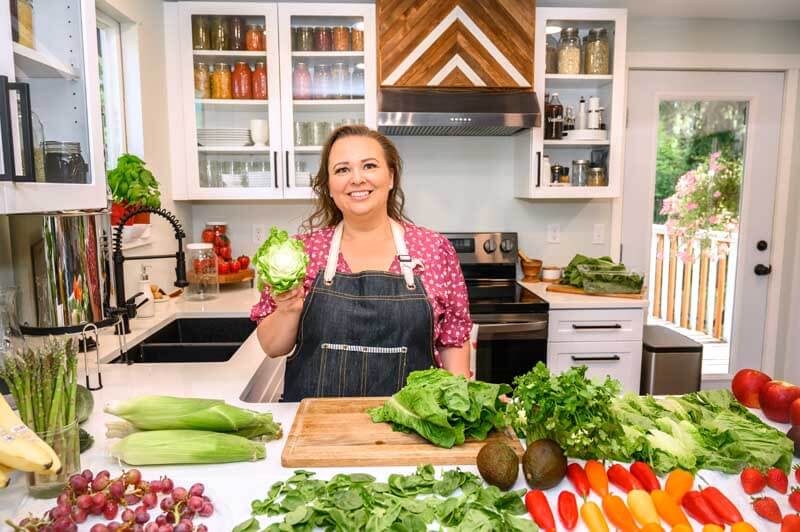 Amy Cross in her kitchen with a spread of fresh produce on the counter in front of her. All this produce from one shopping trips will last us a whole month!