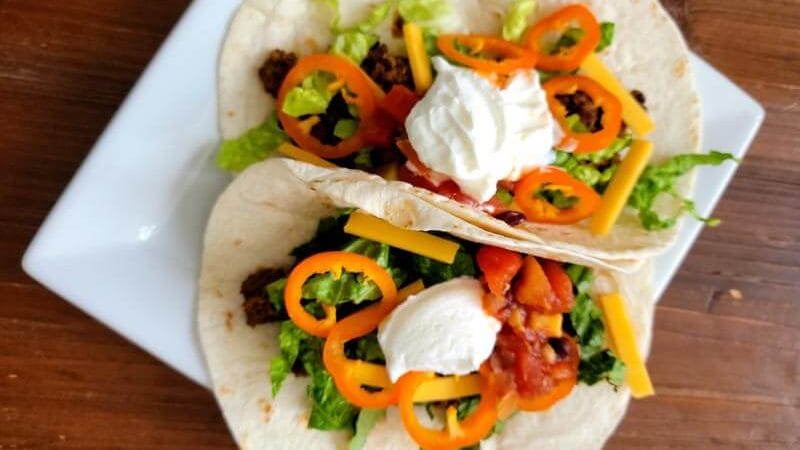 Two finished soft shell tacos with sliced peppers, shredded cheese and a dallop of sour cream on a square white plate.