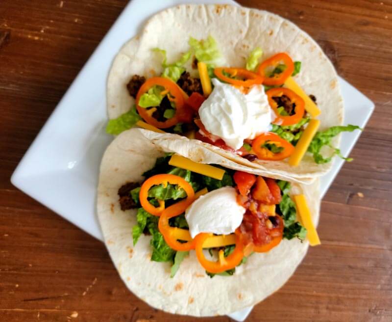 Two finished soft shell tacos with sliced peppers, shredded cheese and a dallop of sour cream on a square white plate.