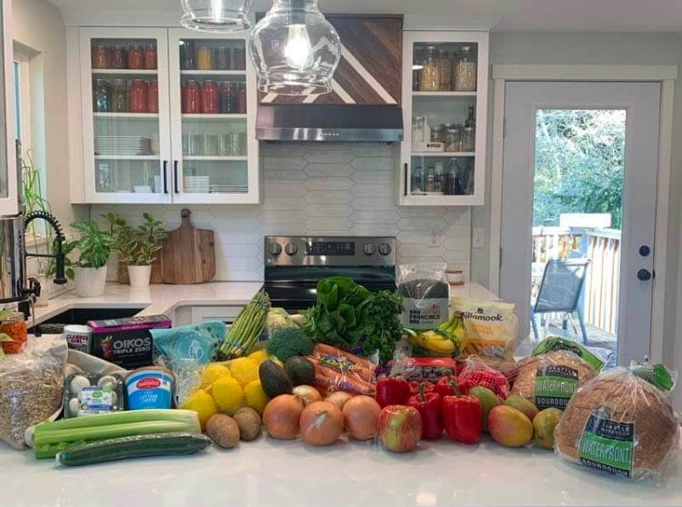 Full spread of groceries on Amy's kitchen counter to include: celery, cucumber, grains, mushrooms, potatoes, lemons, onions, carrots, greens, bananas, peppers, mango, bread, and more!