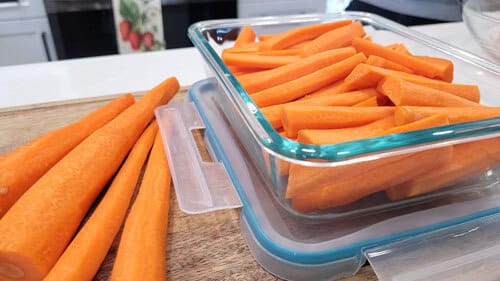 Whole carrots and cut carrots stored in an airtight glass container, sitting on a wooden cutting board.
