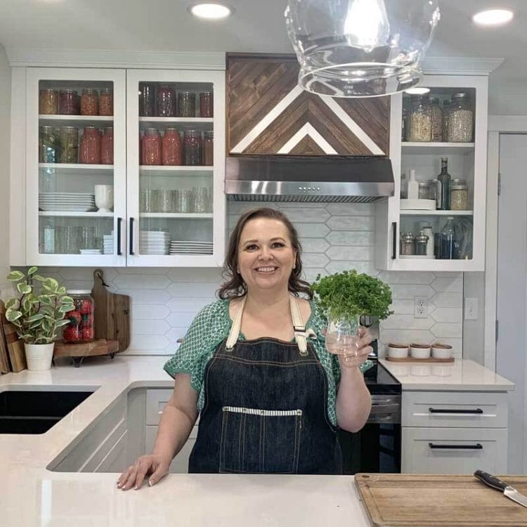 Amy Cross in her kitchen holding fresh herbs in a glass jar with water.