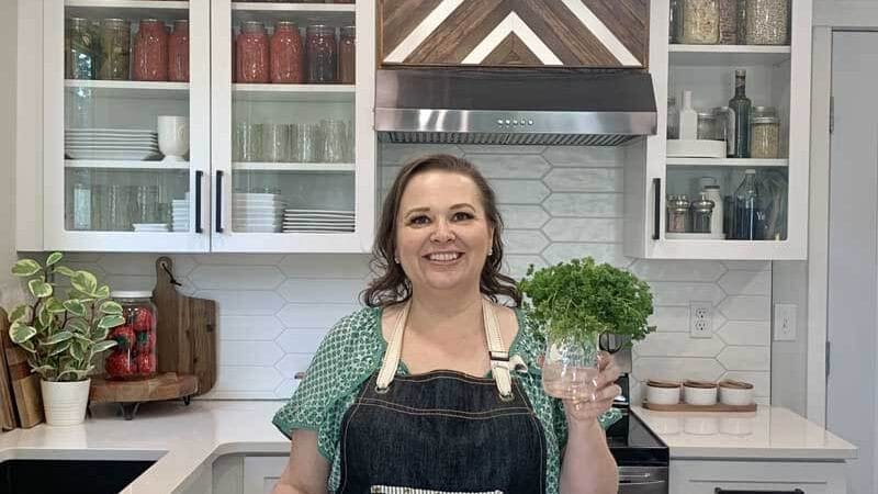 Amy Cross in her kitchen holding fresh herbs in a glass jar with water.