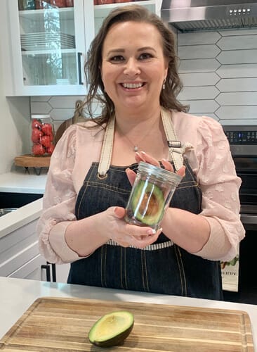 Amy Cross in her kitchen holding a glass mason jar with a cut avocado in it.