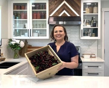 Amy Cross in her kitchen holding a wooden crate full of fresh cherries.