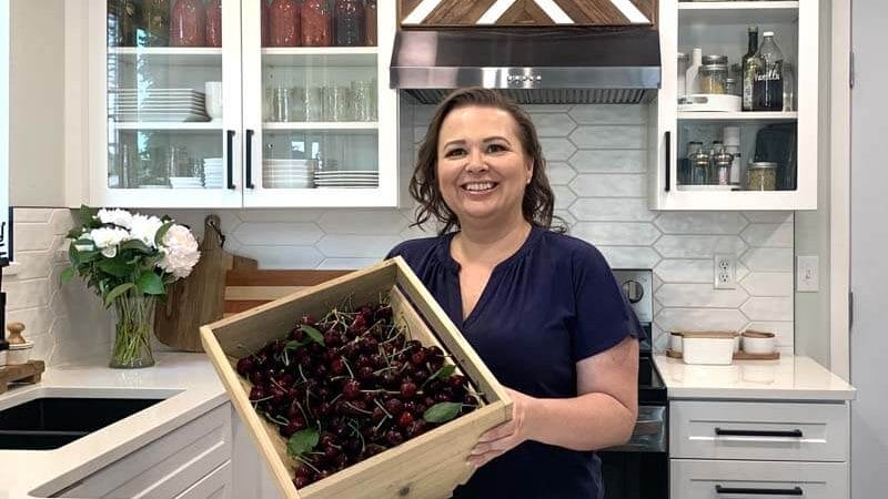Amy Cross in her kitchen holding a wooden crate full of fresh cherries.