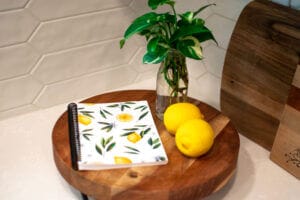 The Lemon Floral The Cross Legacy Grocery Guide and Meal Planner displayed on a wooden surface with a lemon and Pothos cuttings.