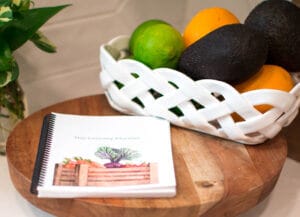 The Rustic Vegetable Box The Cross Legacy Grocery Guide and Meal Planner displayed on a wooden surface with fresh citrus fruits and avocados in a white ceramic woven basket.