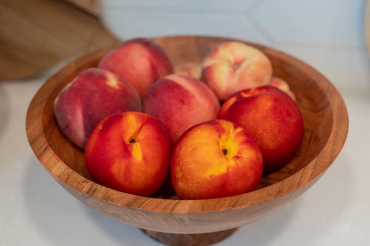 Wooden bowl of fresh peaches on a white counter.