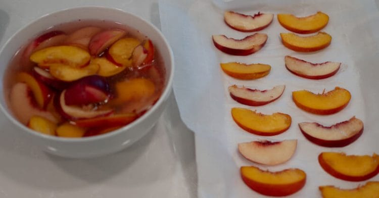 Prepping peach slices for freezing. Some slices in a bowl with lemon juice and other slices on parchment paper in a single layer.