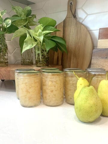 8 jars of canned pears and 3 whole pears on a white counter.