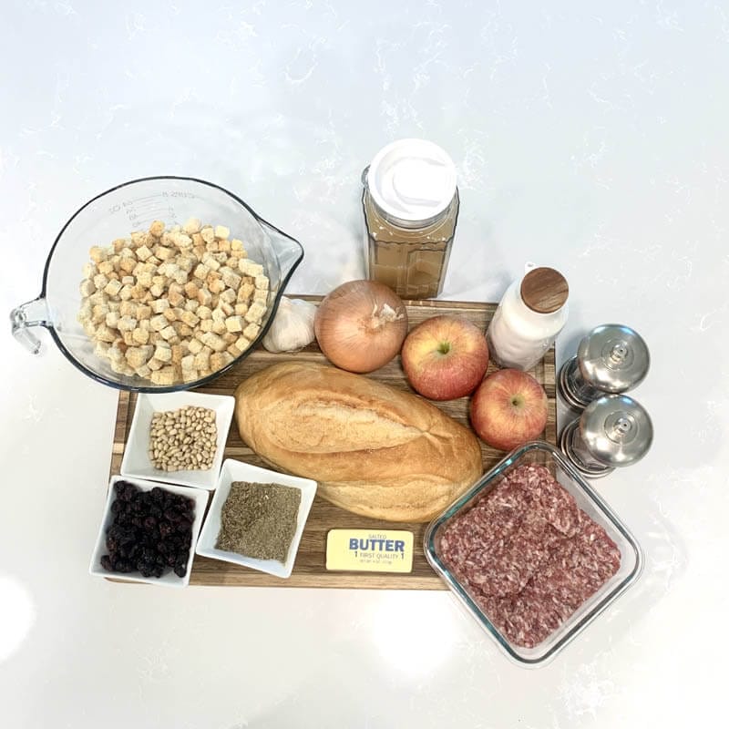 Ingredients to make homemade stuffing laid out on the counter. Bread cubes, garlic clove, onion, two apples, spices, butter, ground sausage.