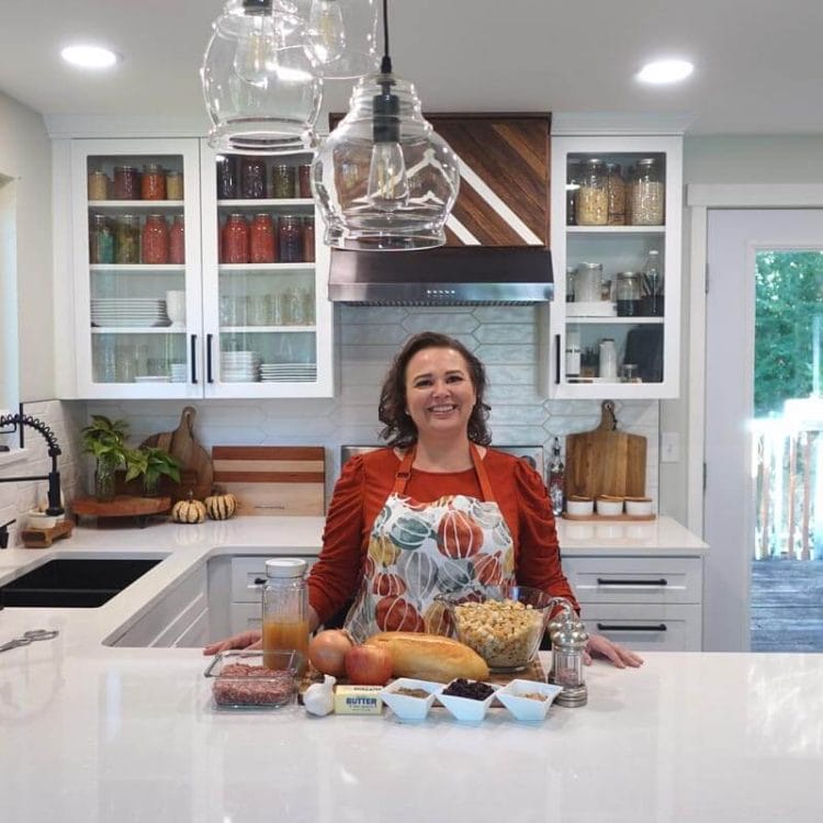 Amy Cross in her kitchen with a fall apron on. Stuffing ingredients are laid out on the counter in front of her.